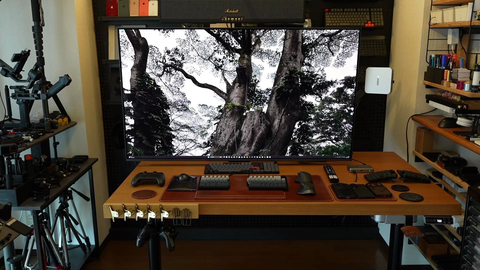 木製デスクに設置した55型テレビ。画面いっぱいに雪をかぶった大木の風景を表示し、手前にキーボードとマウス、左右に工具や機材の棚が並ぶ