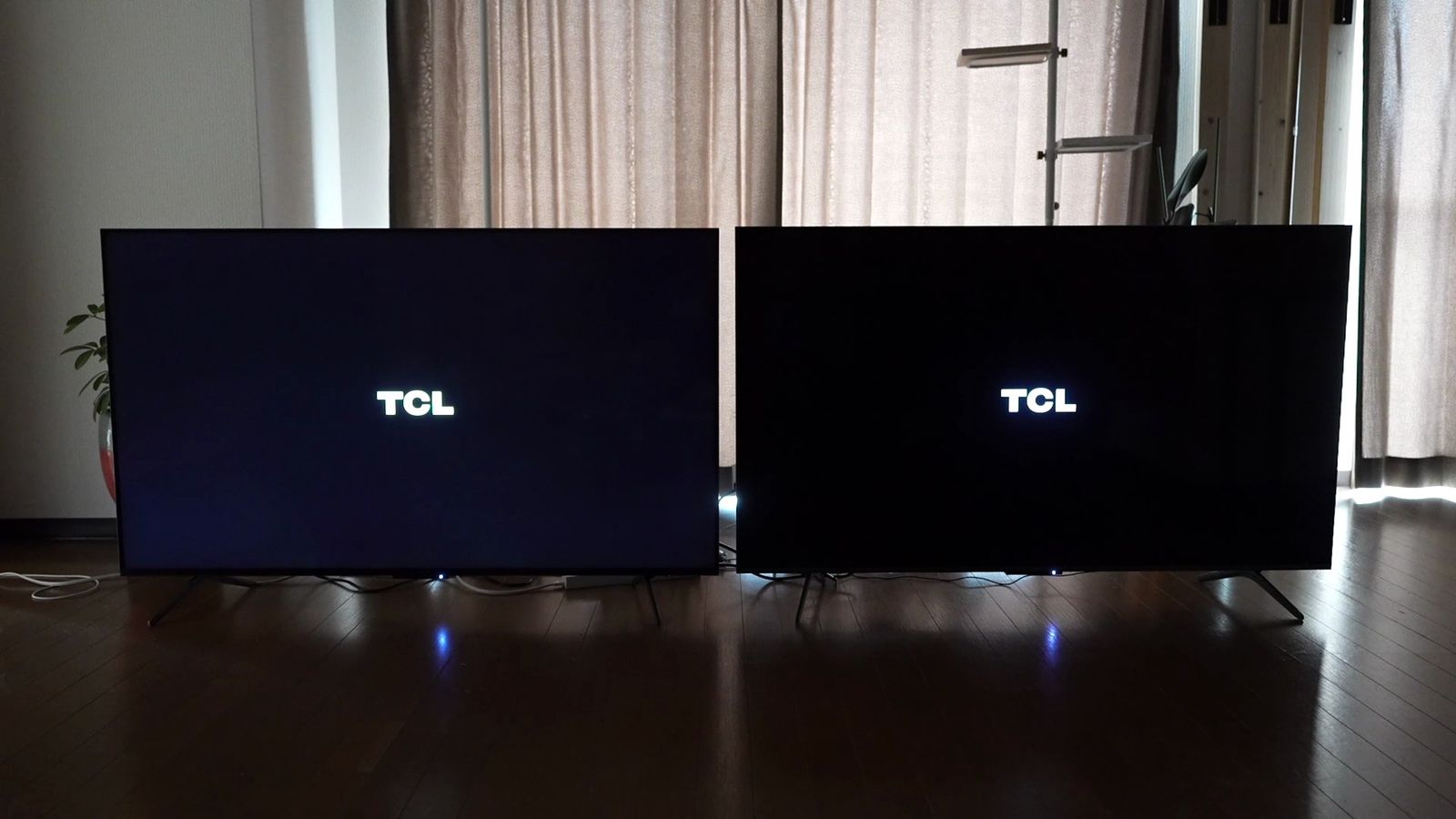 暗めの部屋に並べた2台のTCLテレビ。どちらも起動時のTCLロゴを黒背景に表示し、画面の黒の沈み方を見比べている