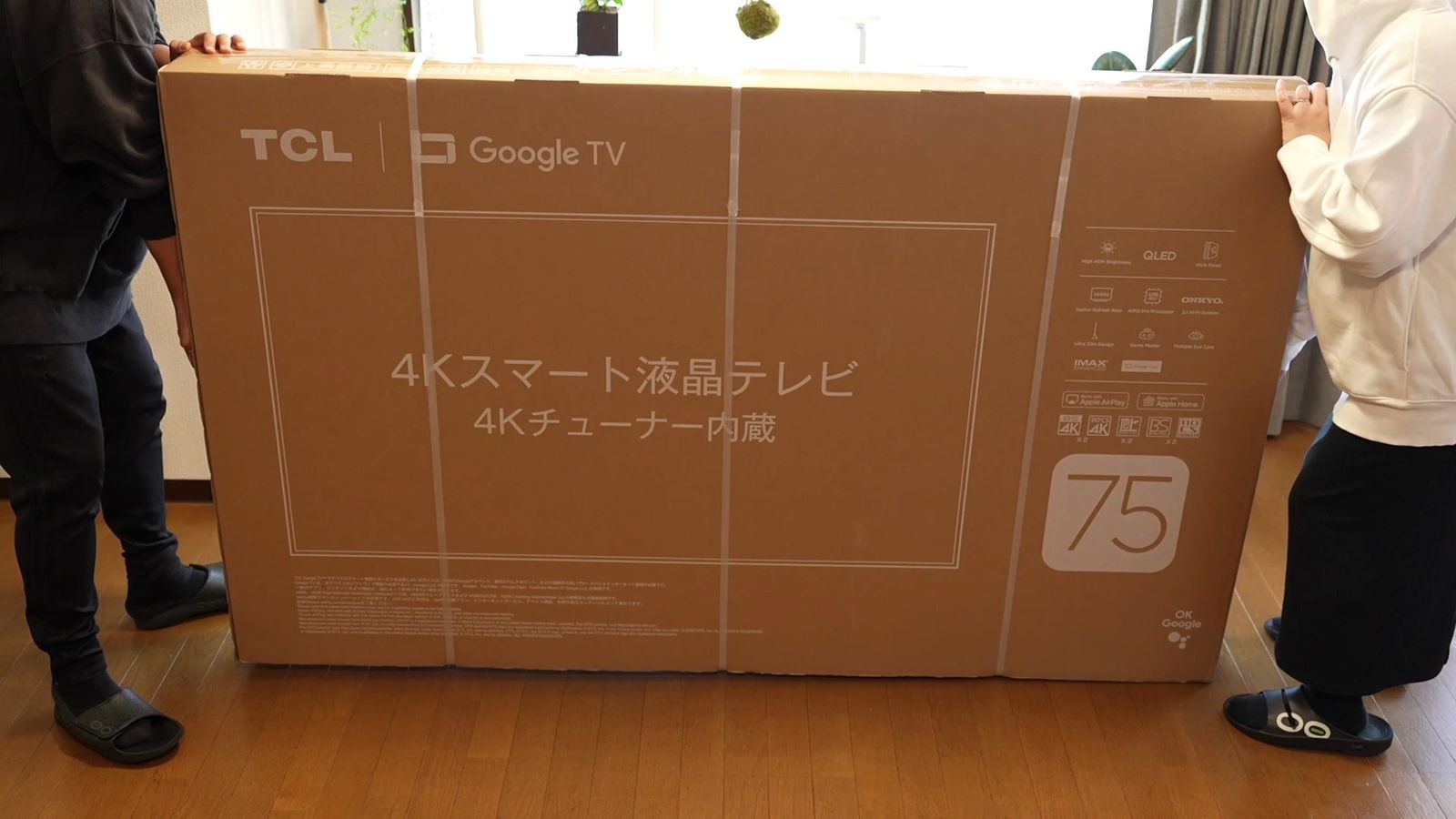 TCLとGoogle TVのロゴ、75の表示がある4Kスマート液晶テレビの外箱を2人で運んでいるところ