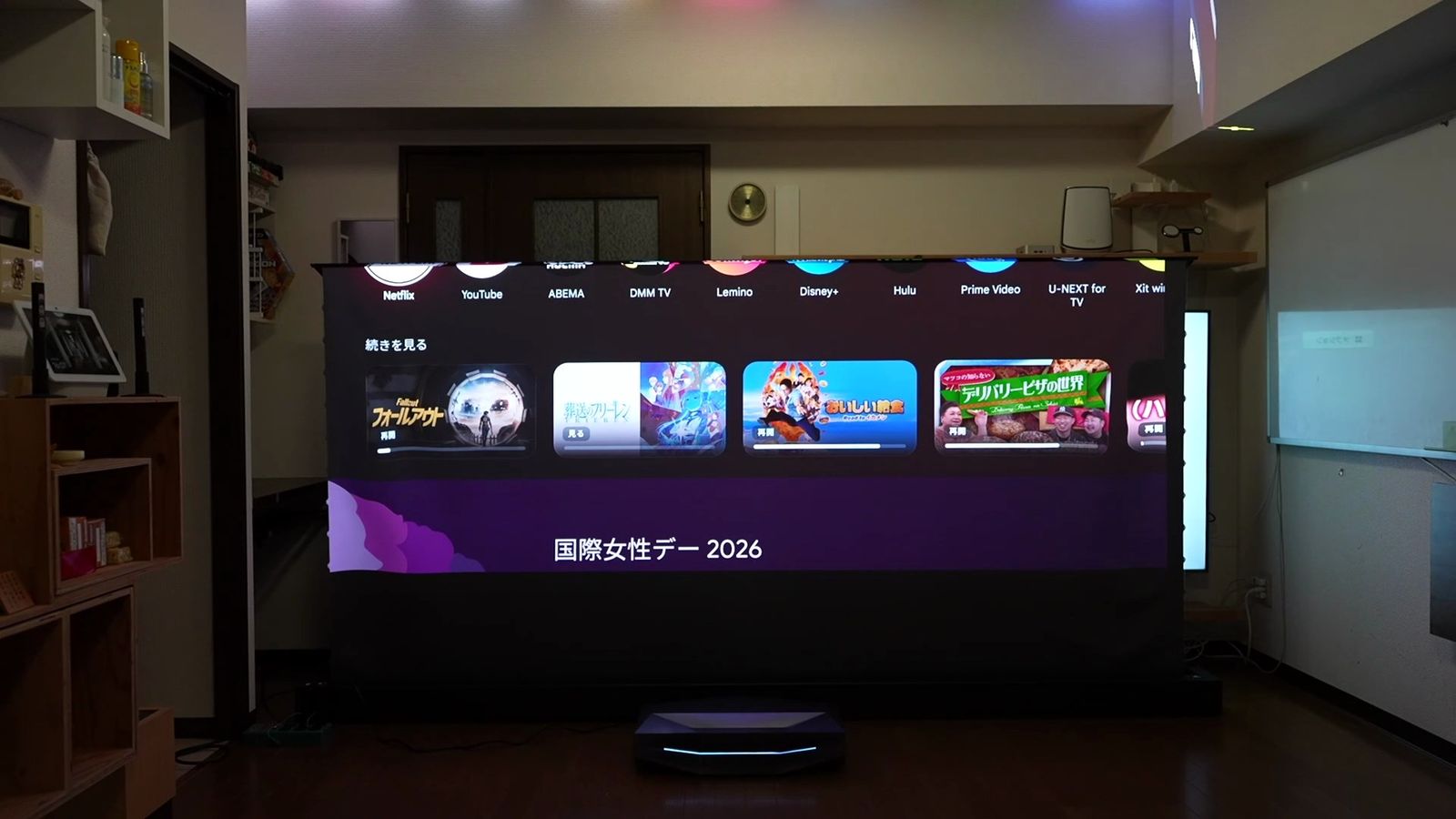 明るいリビングで、床置きのスクリーンにGoogle TVのホーム画面を投影したところ