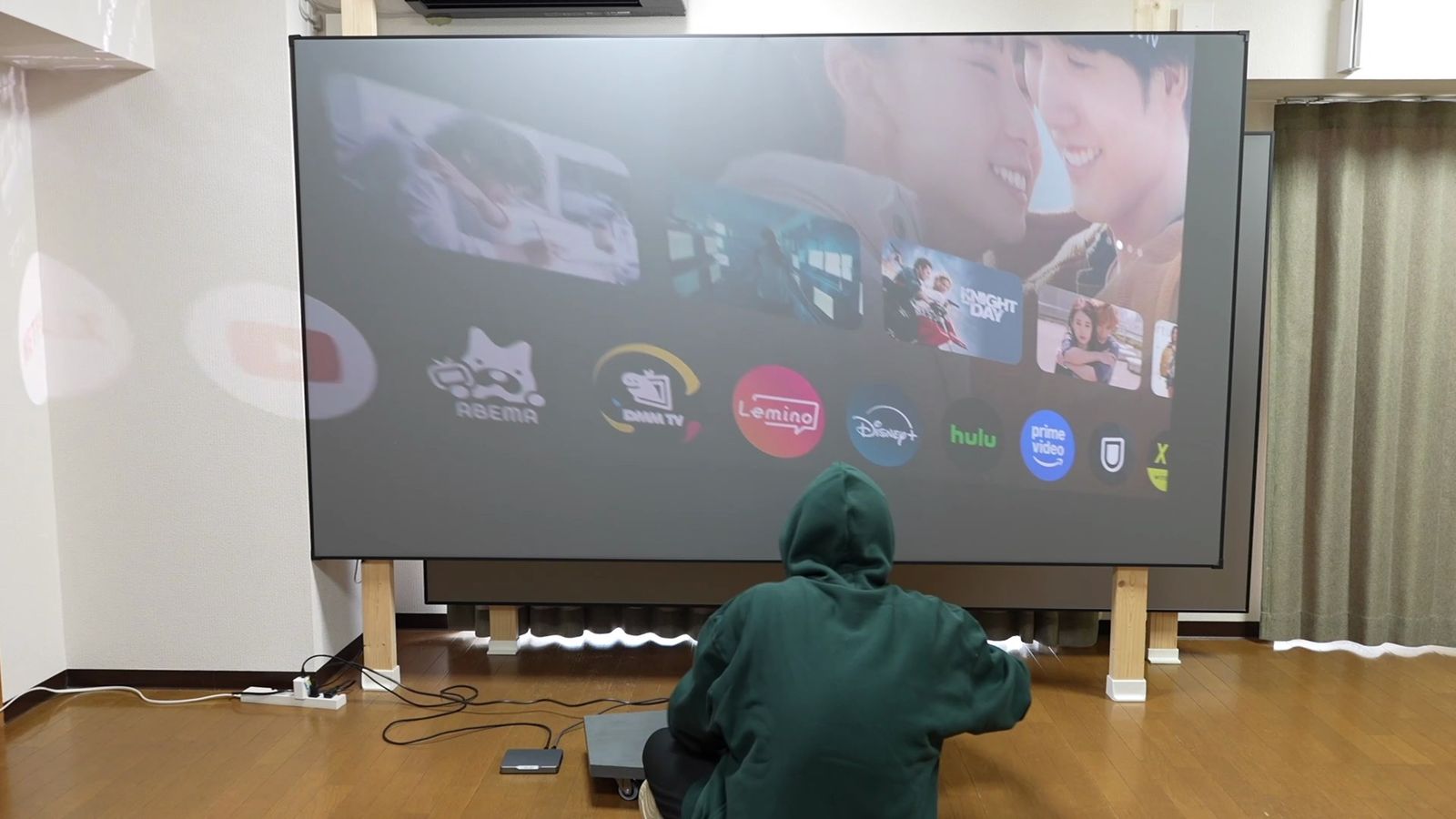 明るい部屋で、柱に掛けたグレーのスクリーンにGoogle TVのホーム画面を投影し、その前に座っているところ